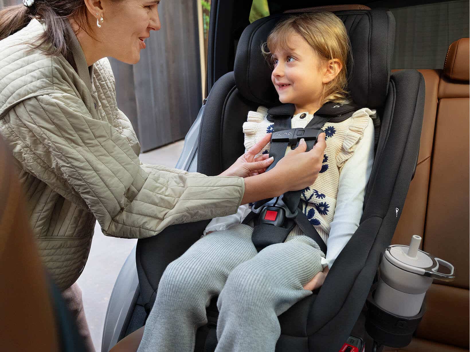 Parent buckling a smiling girl into the black Nuna RAVA next car seat with a sippy cup in the cup holder